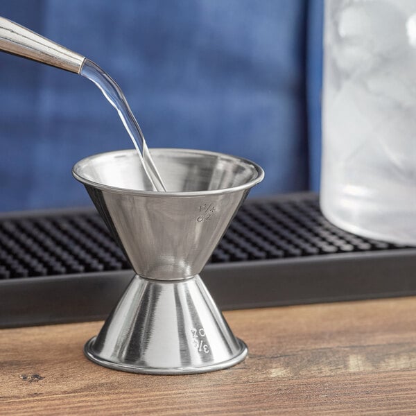 A silver metal Acopa Classic Jigger pouring liquid into a glass.