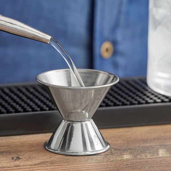 A person using an Acopa stainless steel jigger to pour a drink into a metal funnel.