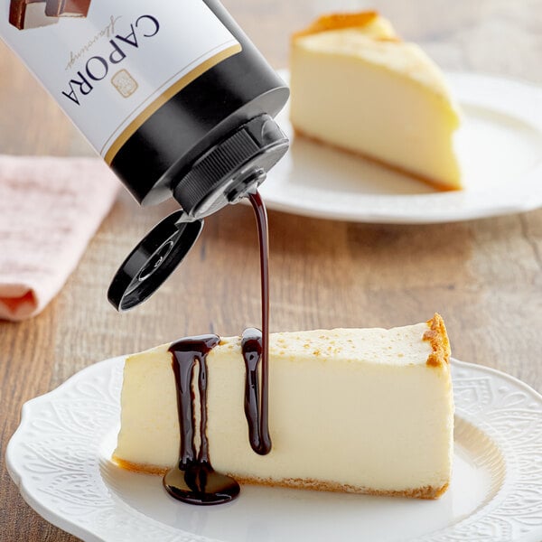 A Capora bottle pouring chocolate sauce onto a slice of cheesecake.