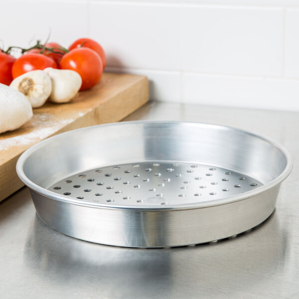 An American Metalcraft heavy weight aluminum pizza pan with holes on it next to tomatoes on a cutting board.