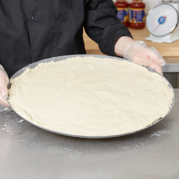 A person holding a round white pizza dough in an American Metalcraft Super Perforated Hard Coat Anodized Aluminum Pizza Pan.