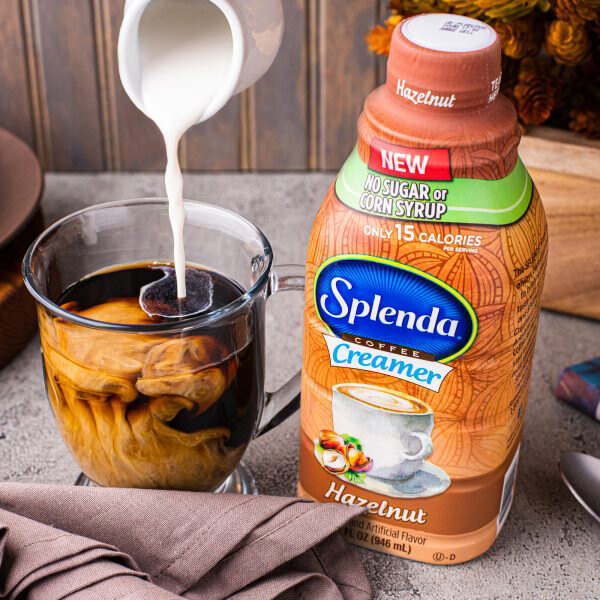 A close-up of a Splenda Sugar-Free Hazelnut Coffee Creamer container on a table with a glass of coffee being poured into.
