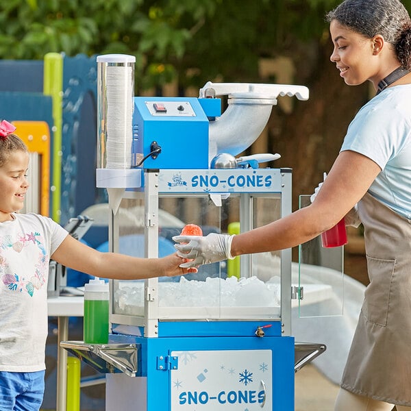A woman wearing gloves uses a Carnival King snow cone machine to make a green snow cone for a girl.