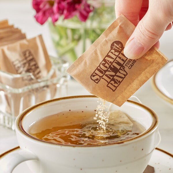 A hand pouring a Sugar In The Raw packet into a cup of tea.