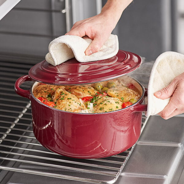 A person using a white towel to put a red Valor enameled cast iron Dutch oven with a red lid into an oven.