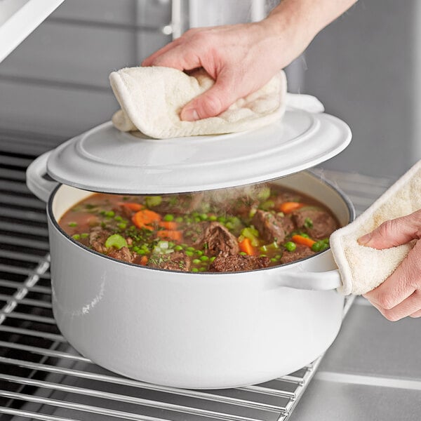 A person's hand holding a white towel wiping the Valor enameled cast iron Dutch oven filled with soup.
