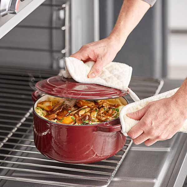 A person using a white towel to remove the lid from a red Valor enameled cast iron Dutch oven.