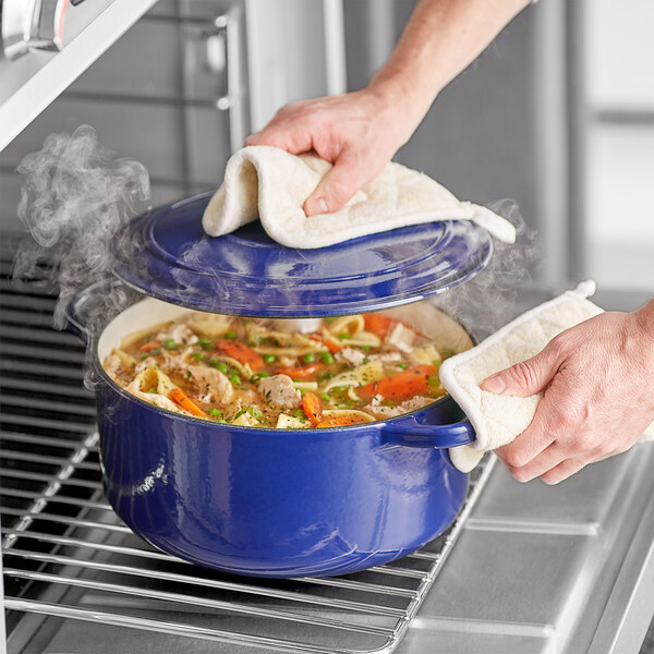 A hand holding a blue Valor enameled cast iron Dutch oven filled with noodle soup over a white towel.