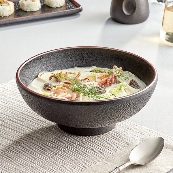 A black Acopa Heika stoneware bowl filled with soup on a table with a spoon.