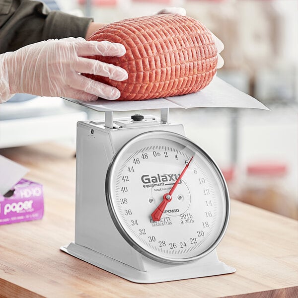 A person weighing meat on a white Galaxy mechanical portion scale with a red needle.