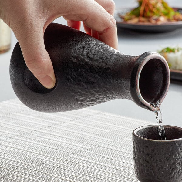 A hand pouring liquid into a small black Acopa stoneware sake bottle.