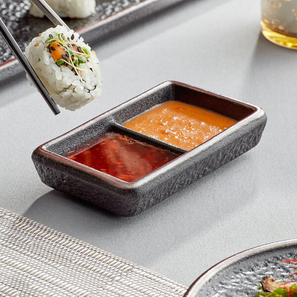 A black rectangular Acopa stoneware dish with 2 compartments holding sushi and sauce.