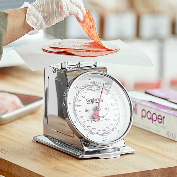 A person weighing meat on a Galaxy 5 lb. mechanical portion scale on a wood surface.