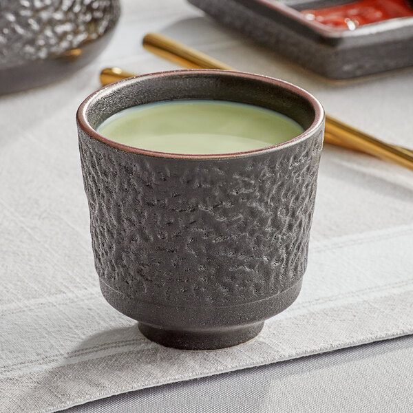 A black Acopa Heika stoneware tea cup filled with green liquid on a table.