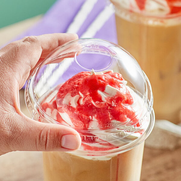 A hand holding a Choice clear plastic dome lid on a plastic cup with a red and white topping.