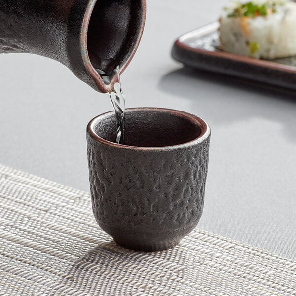 A pitcher pouring sake into a black Acopa stoneware cup.
