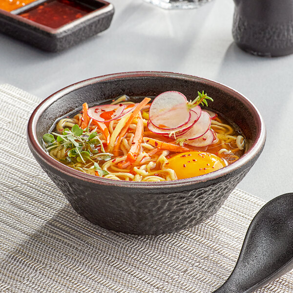 A black Acopa stoneware bowl filled with ramen and vegetables with a black spoon.