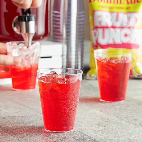 A hand pours DominAde Fruit Punch Drink Mix into a glass of red liquid.
