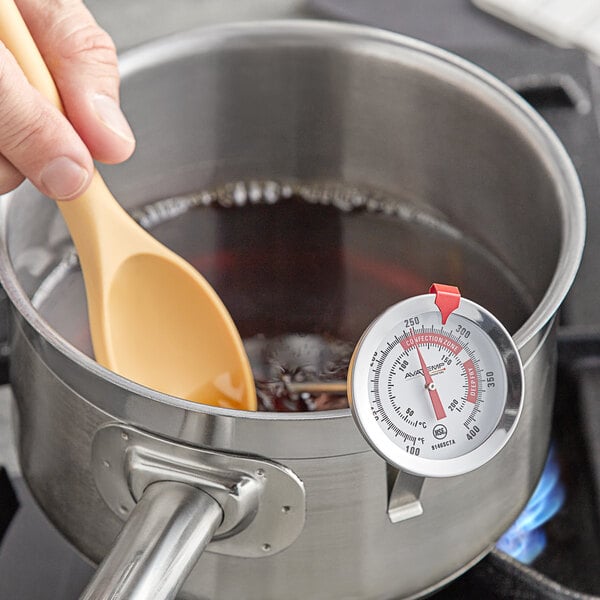 A hand holding an AvaTemp candy thermometer in a pot of liquid.