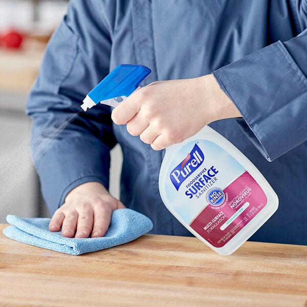 A person in blue clothes using Purell Foodservice Surface Sanitizer to spray a table.