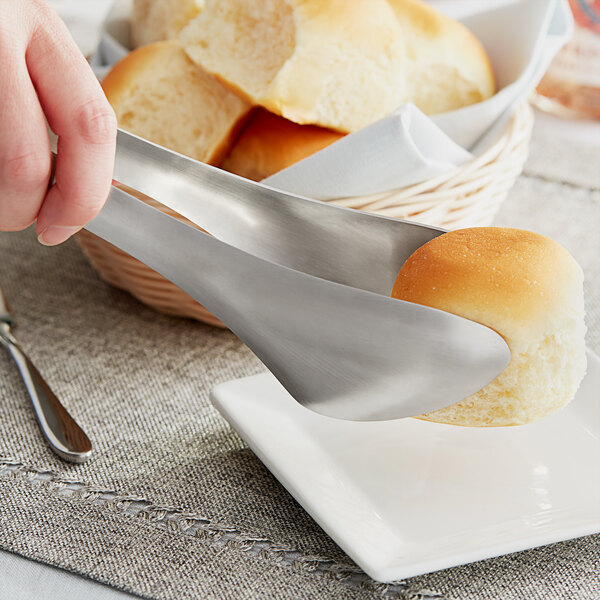 A person using American Metalcraft stainless steel tongs to pick up a bread roll.