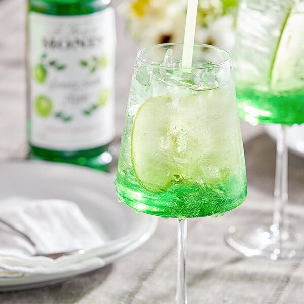 A glass of green liquid with a straw sitting on a table with a white bottle of Monin Premium Granny Smith Apple Fruit Syrup.