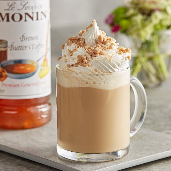 A glass mug of Monin brown butter toffee flavoring syrup in coffee with whipped cream and nuts.