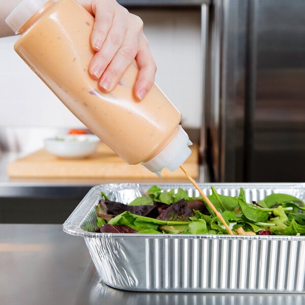 A person using a FIFO Innovations squeeze bottle to pour sauce on a salad in a foil pan on a counter.
