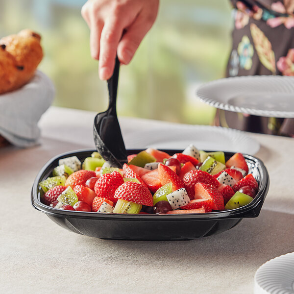 A person using a fork to eat fruit from a Visions black plastic serving bowl on a table.