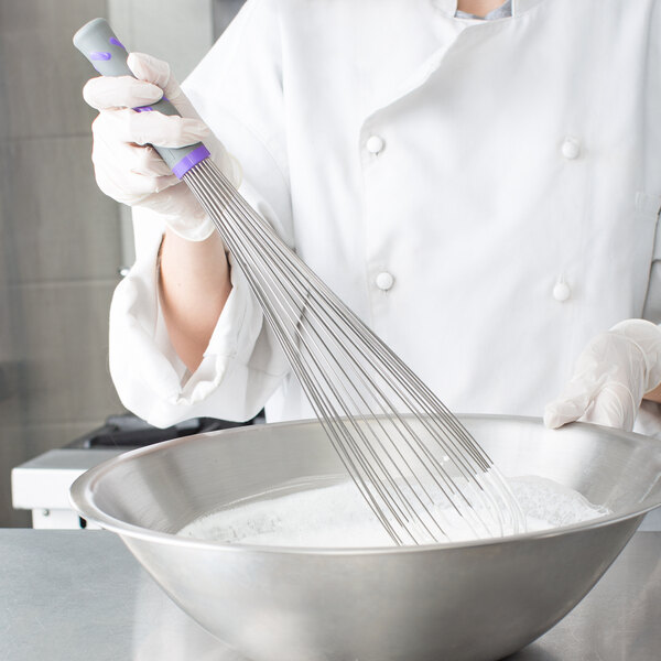 A person in a white coat using a Vollrath stainless steel piano whisk to mix ingredients in a bowl.