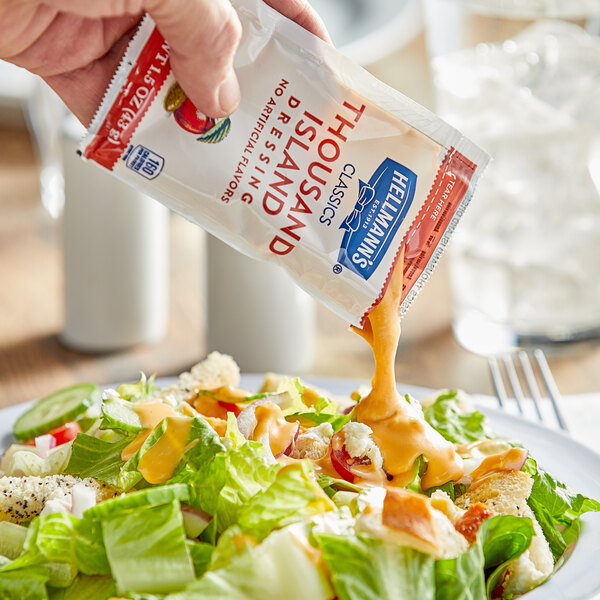A hand pouring Hellmann's Thousand Island dressing from a packet onto a salad.