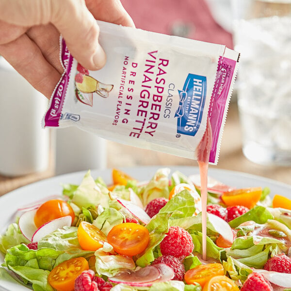 A hand pouring a Hellmann's Raspberry Vinaigrette dressing packet over a salad on a table.