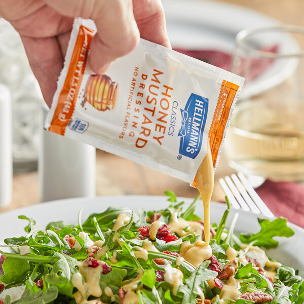 A hand pouring Hellmann's Honey Mustard dressing from a packet onto a salad.