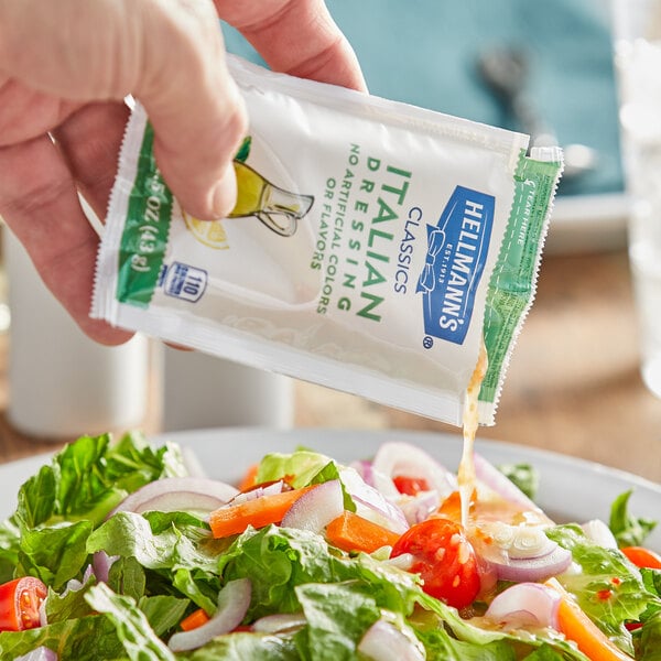 A hand pouring a Hellmann's Zesty Italian Dressing packet over a salad on a table in a salad bar.