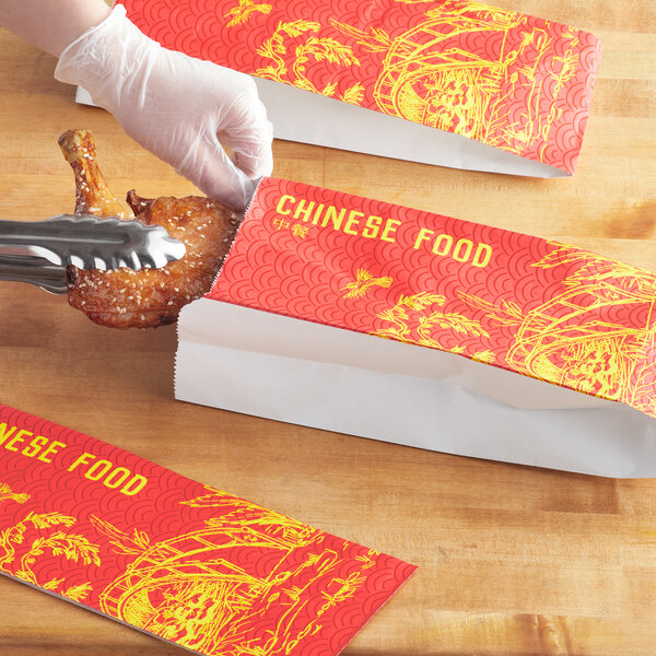 A person cutting a piece of chicken in a red and yellow Emperor's Select Chinese food bag.