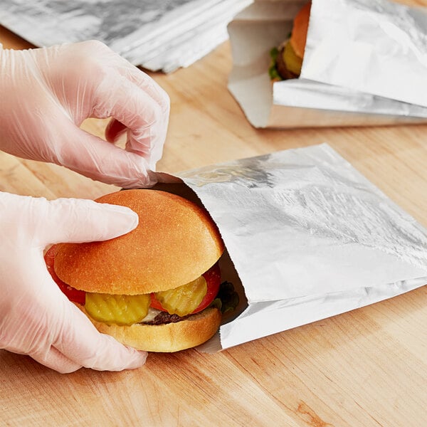 A person wearing gloves puts a hamburger in a Carnival King foil sandwich bag.