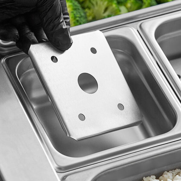 A hand holding a Choice 1/6 size stainless steel false bottom over a tray of food.