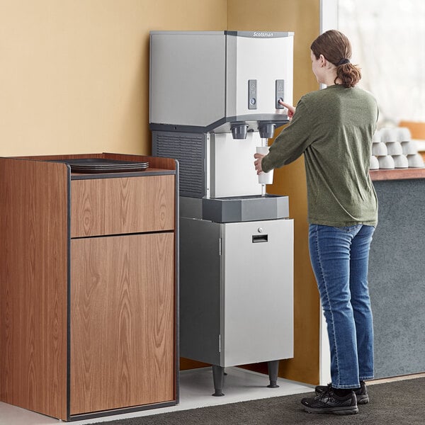 A woman using a Scotsman Meridian ice and water dispenser on a white cabinet stand.