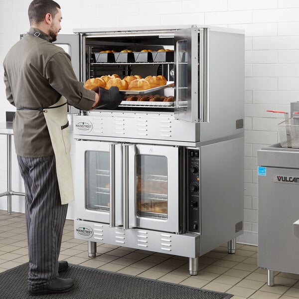A man standing in a commercial kitchen with a Main Street Equipment double deck convection oven filled with food.