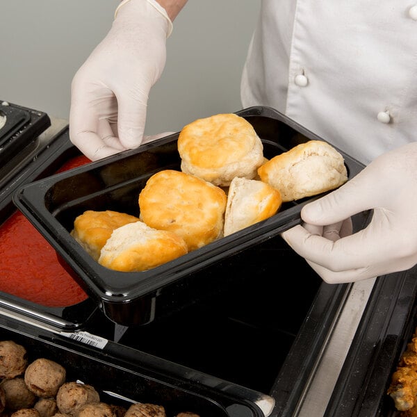 A person in a white chef's uniform holding a Cambro black plastic food pan filled with food.