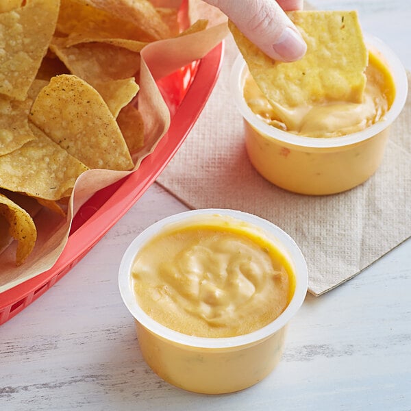 A person holding a tortilla chip and dipping it into a cup of Muy Fresco nacho cheese.