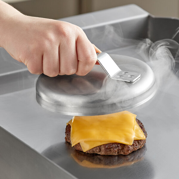 A person using a Vollrath aluminum basting cover to melt cheese on a burger with a metal spatula.