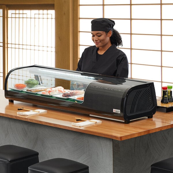A woman standing behind a countertop Emperor's Select refrigerated sushi display case full of sushi.