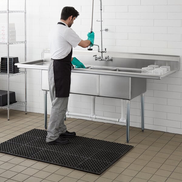 A man in a green apron washing a Steelton stainless steel three compartment sink.