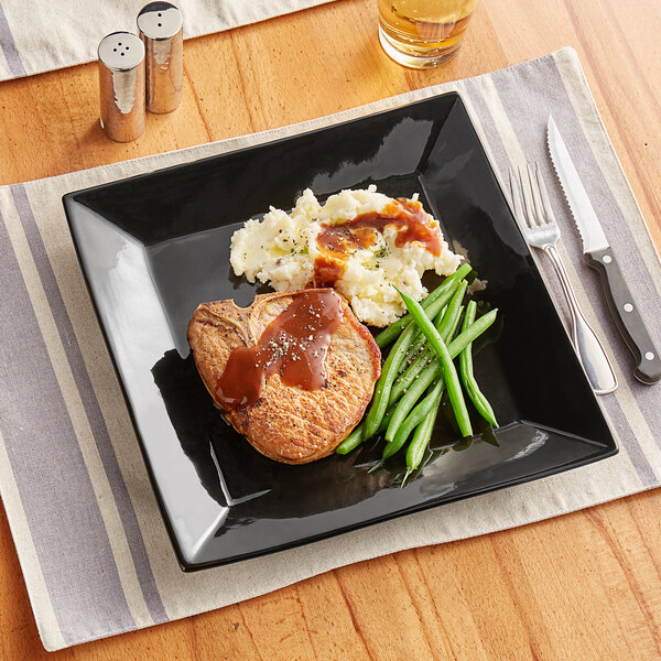A glossy black square stoneware plate with meat, green beans, and mashed potatoes on it.