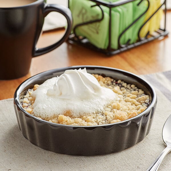 A black Acopa fluted stoneware bowl of dessert with whipped cream on a table with a spoon.