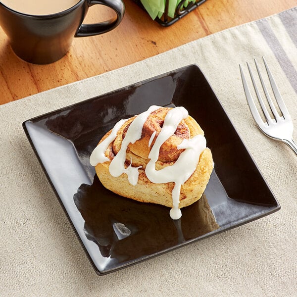 A glossy black Acopa stoneware plate with a cinnamon roll with white icing on top next to a cup of coffee.