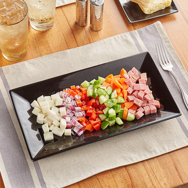 A glossy black Acopa rectangular stoneware platter with sliced meat, cheese, and vegetables on a table.