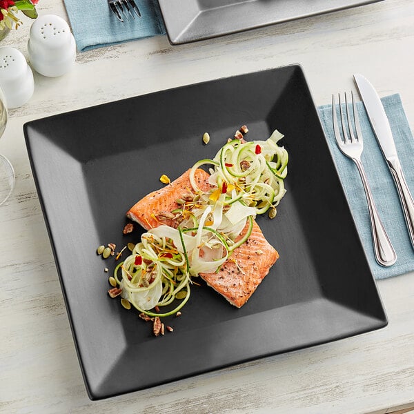 A matte black Acopa stoneware plate with salmon and vegetables on a table