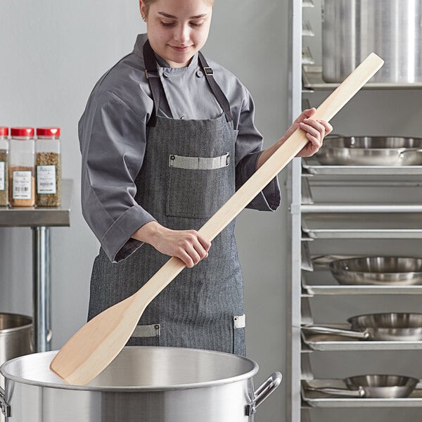 A woman in a chef's uniform using an American Metalcraft wood paddle in a professional kitchen.